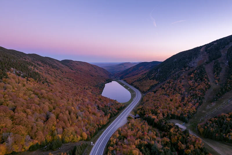 Aerial view of a winding highway through a mountain valley surrounded by autumn foliage and a small lake at dusk