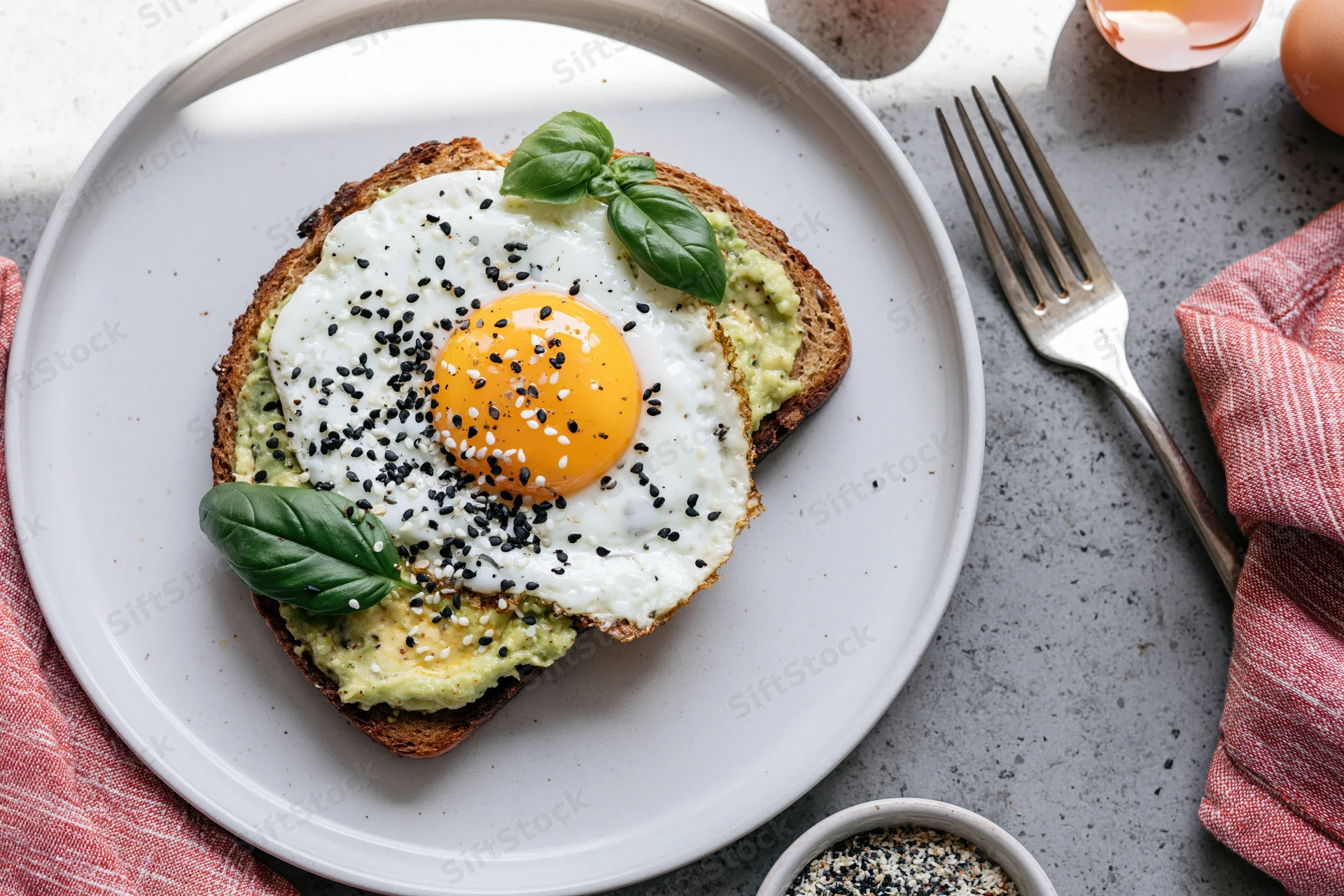 Avocado toast topped with sunny-side-up egg and sesame seeds on a white plate with basil garnish, set on a kitchen table with fork and napkin nearby