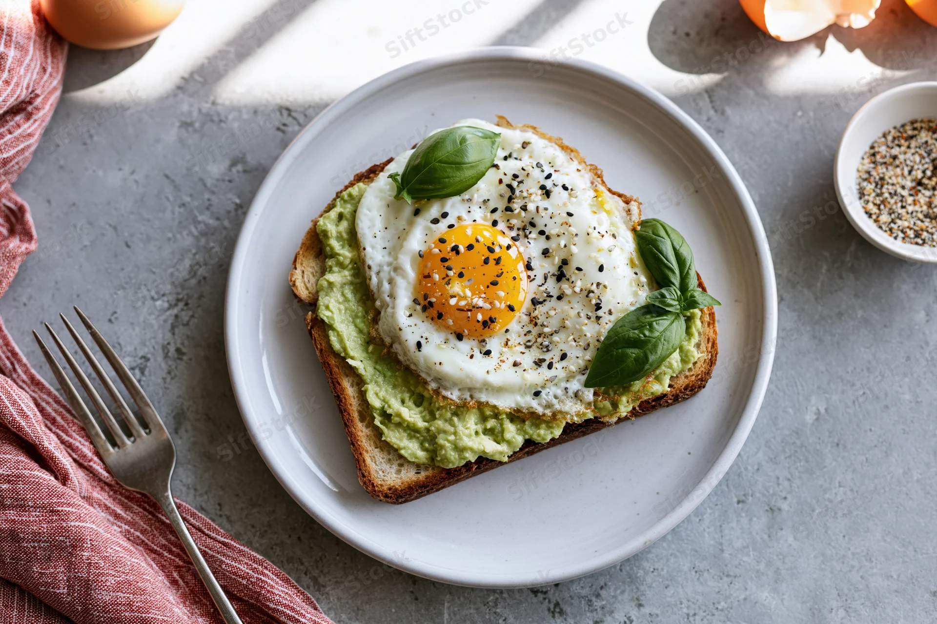 Avocado toast topped with sunny-side-up egg and basil on white plate with fork and napkin on gray surface