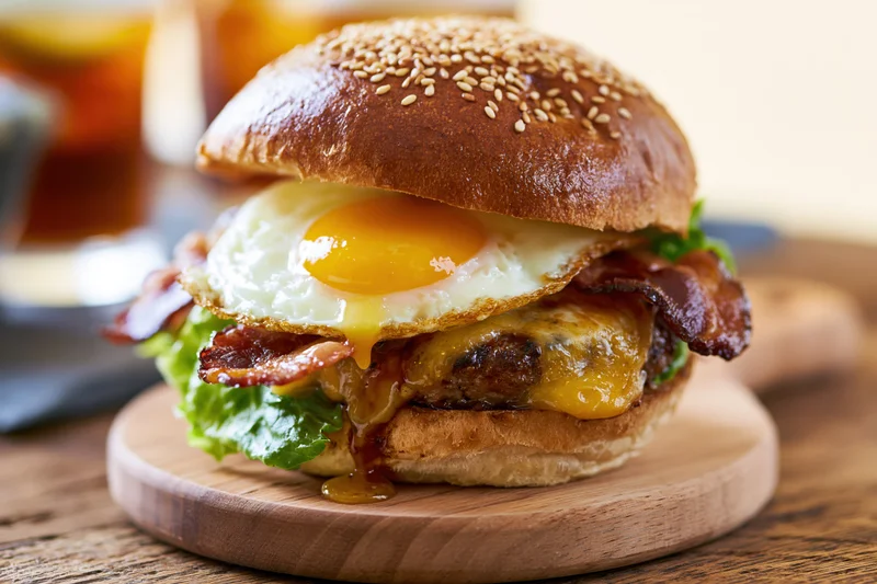 Breakfast burger with fried egg, crispy bacon, melted cheese, and lettuce on a sesame seed bun served on a wooden board
