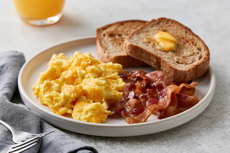 Classic american breakfast plate with scrambled eggs bacon and toast on a white plate with orange juice and napkin on a table