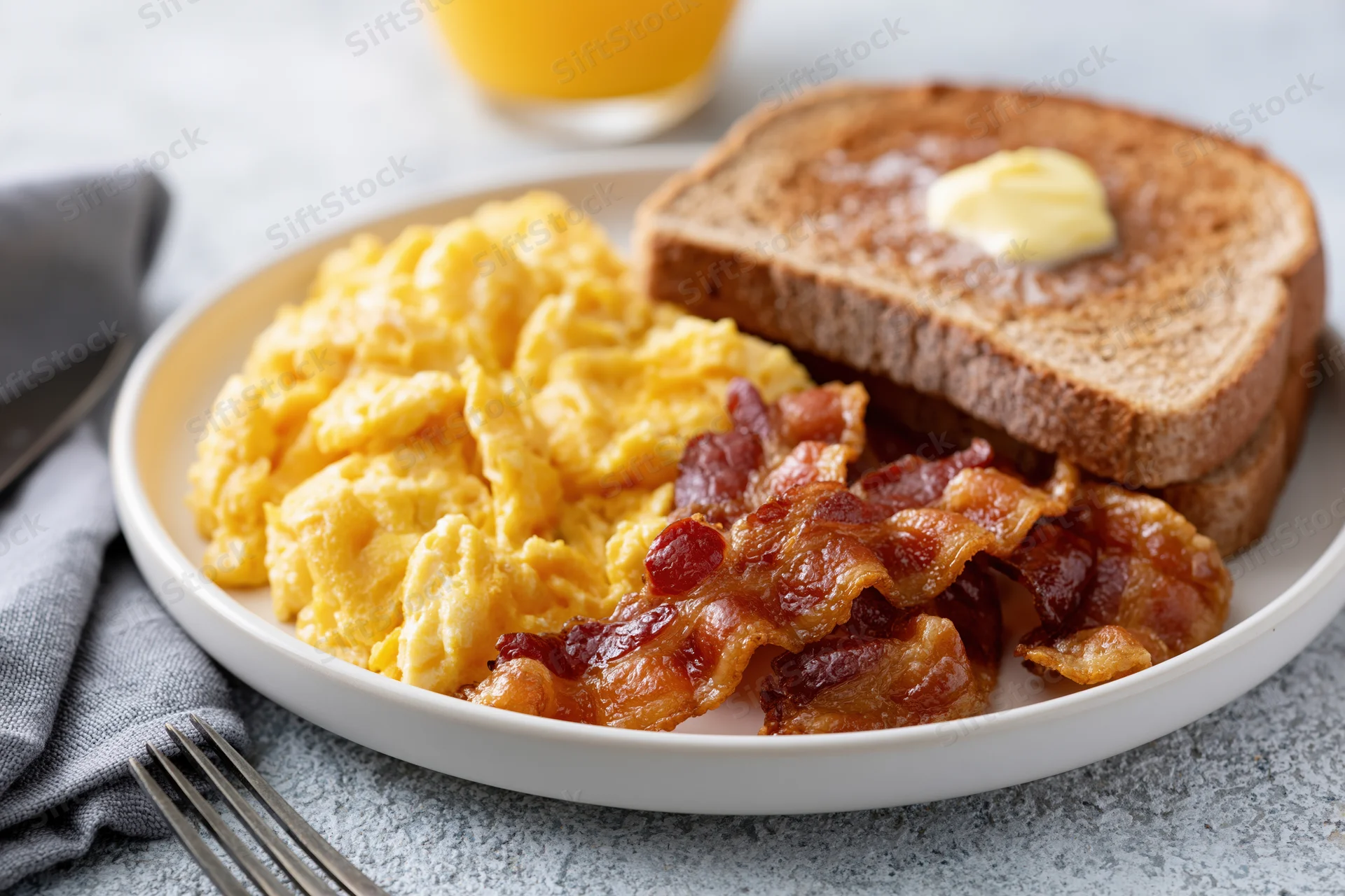 Classic american breakfast plate with scrambled eggs, bacon, and toast on a white plate with orange juice in the background