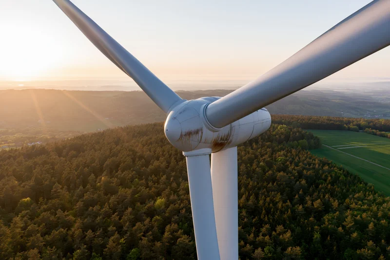 Close aerial view of wind turbine hub and blades above forested landscape at sunset