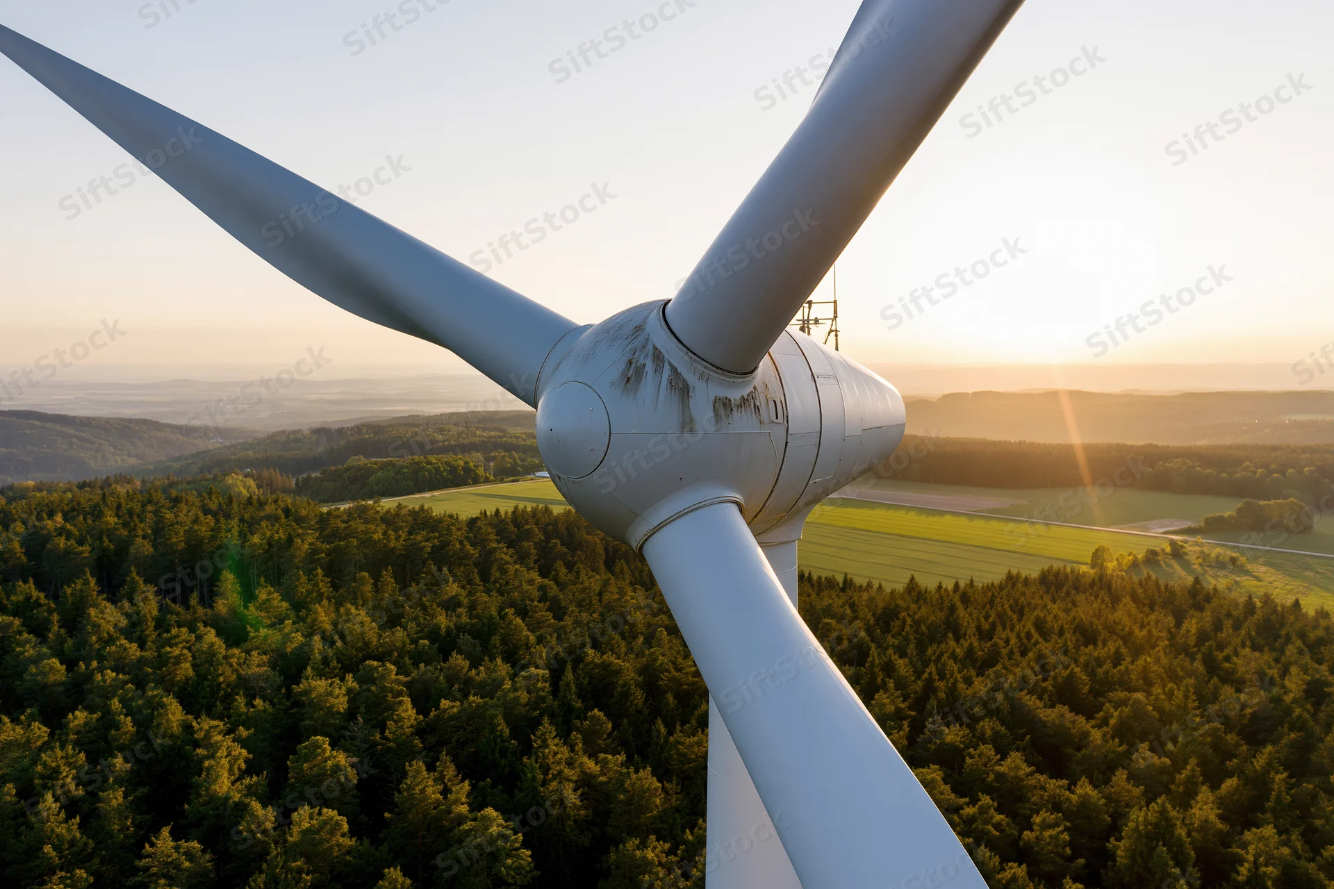 Wind turbine hub and three blades viewed from above over forest and fields at sunset