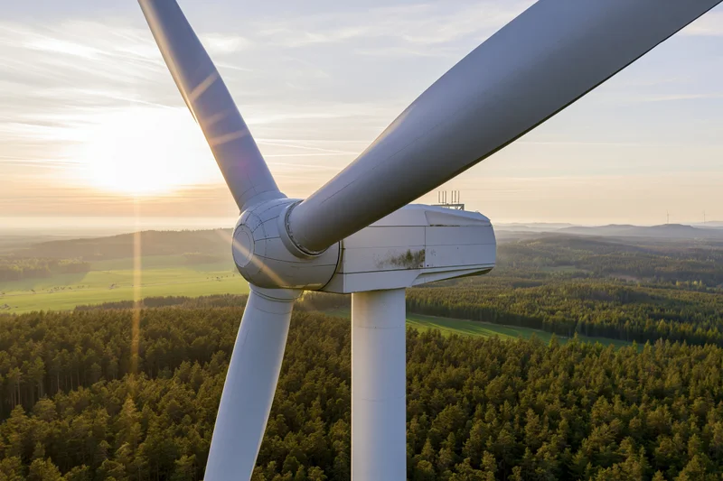 Close aerial view of wind turbine hub and three blades over forested landscape at sunset