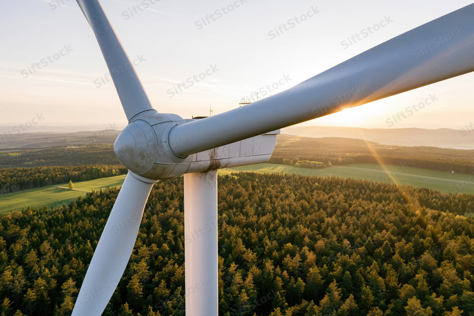 Close aerial view of wind turbine hub and blades over forested countryside at sunset