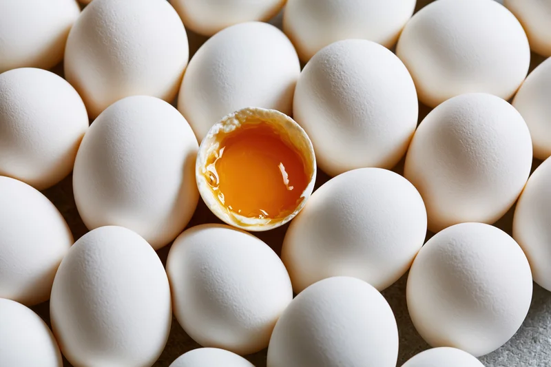 Dozens of whole white eggs packed tightly together with one cracked open revealing yolk in center