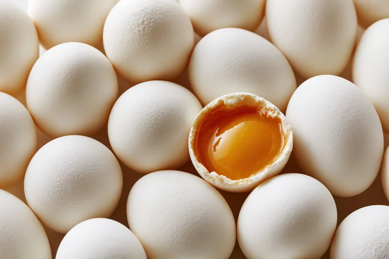 Cracked egg with visible yolk surrounded by whole white eggs in tight arrangement