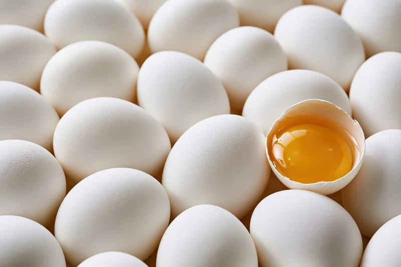 Dozens of whole white eggs packed tightly together with one cracked open revealing yolk