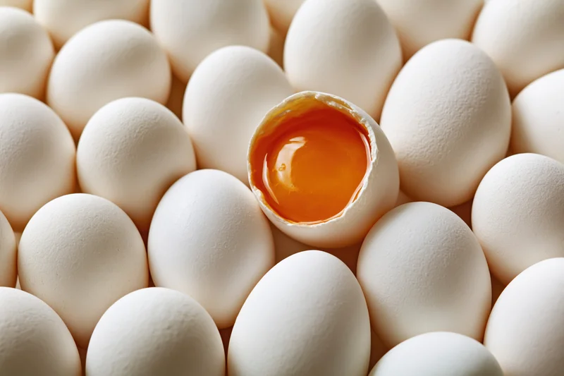 Cracked egg with visible yolk surrounded by whole white eggs in tight arrangement