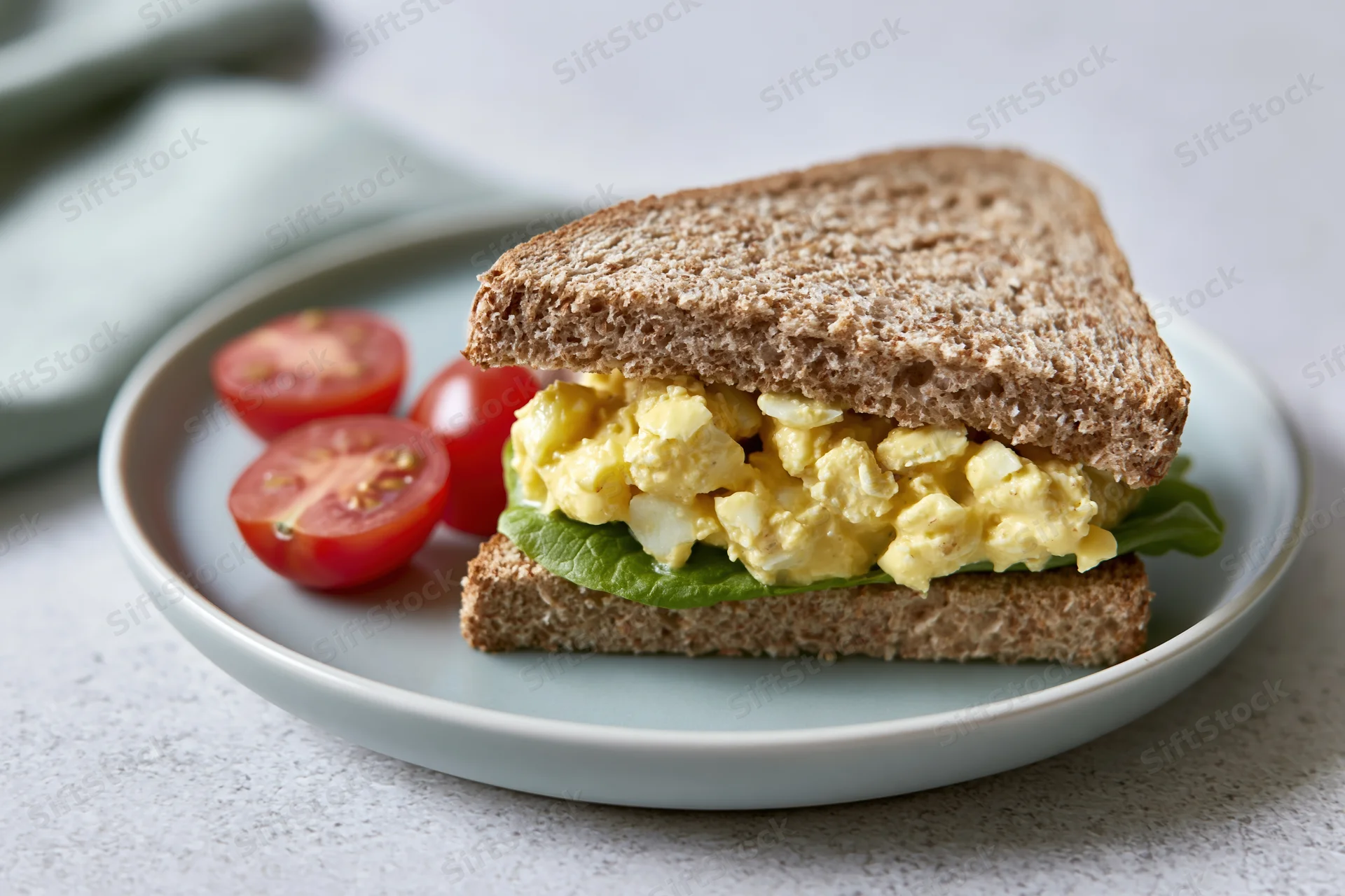 Egg salad sandwich cut diagonally on a light grey ceramic plate with cherry tomatoes and lettuce