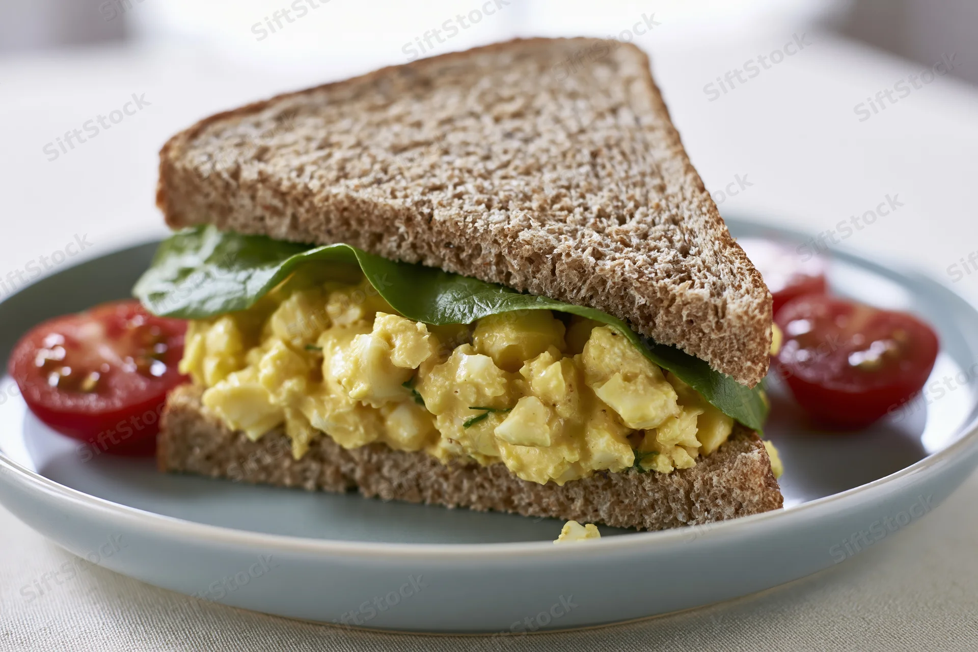 Egg salad sandwich cut diagonally on a light grey ceramic plate with tomato slices and lettuce, served on a white table surface