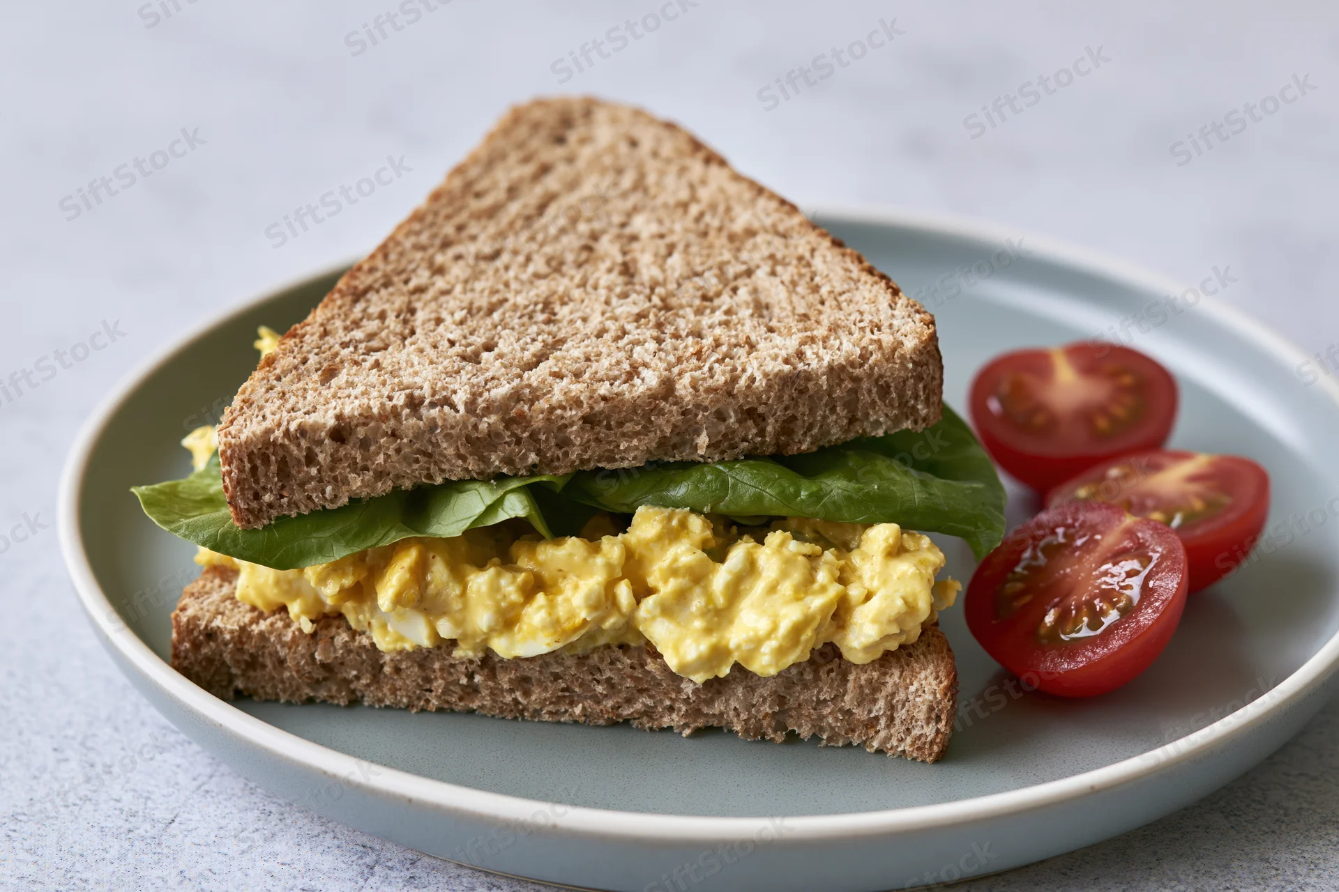 Egg salad sandwich cut diagonally on a light grey ceramic plate with cherry tomato slices beside it