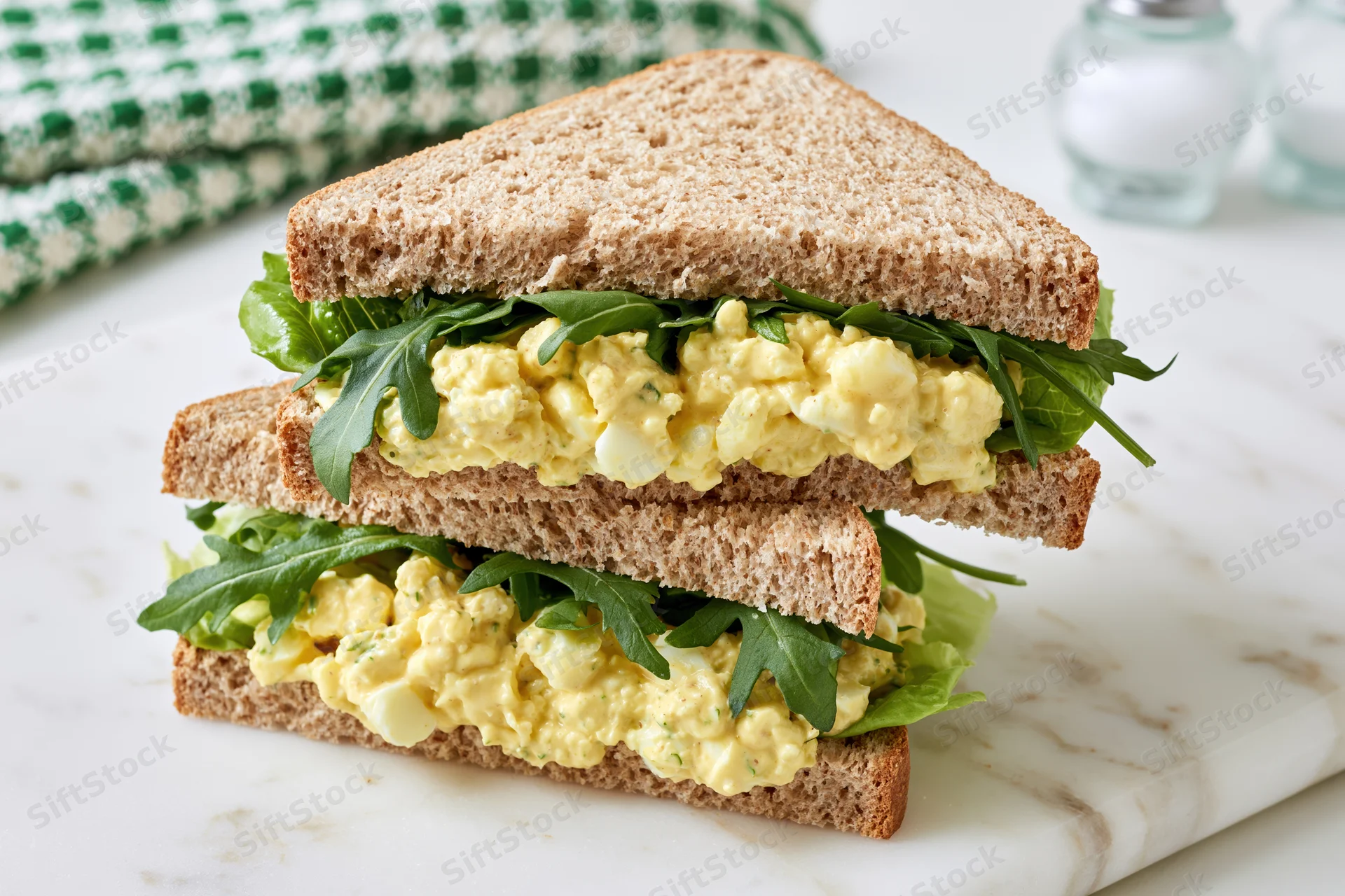 Egg salad sandwich cut in half and stacked on white marble countertop with arugula and lettuce layers