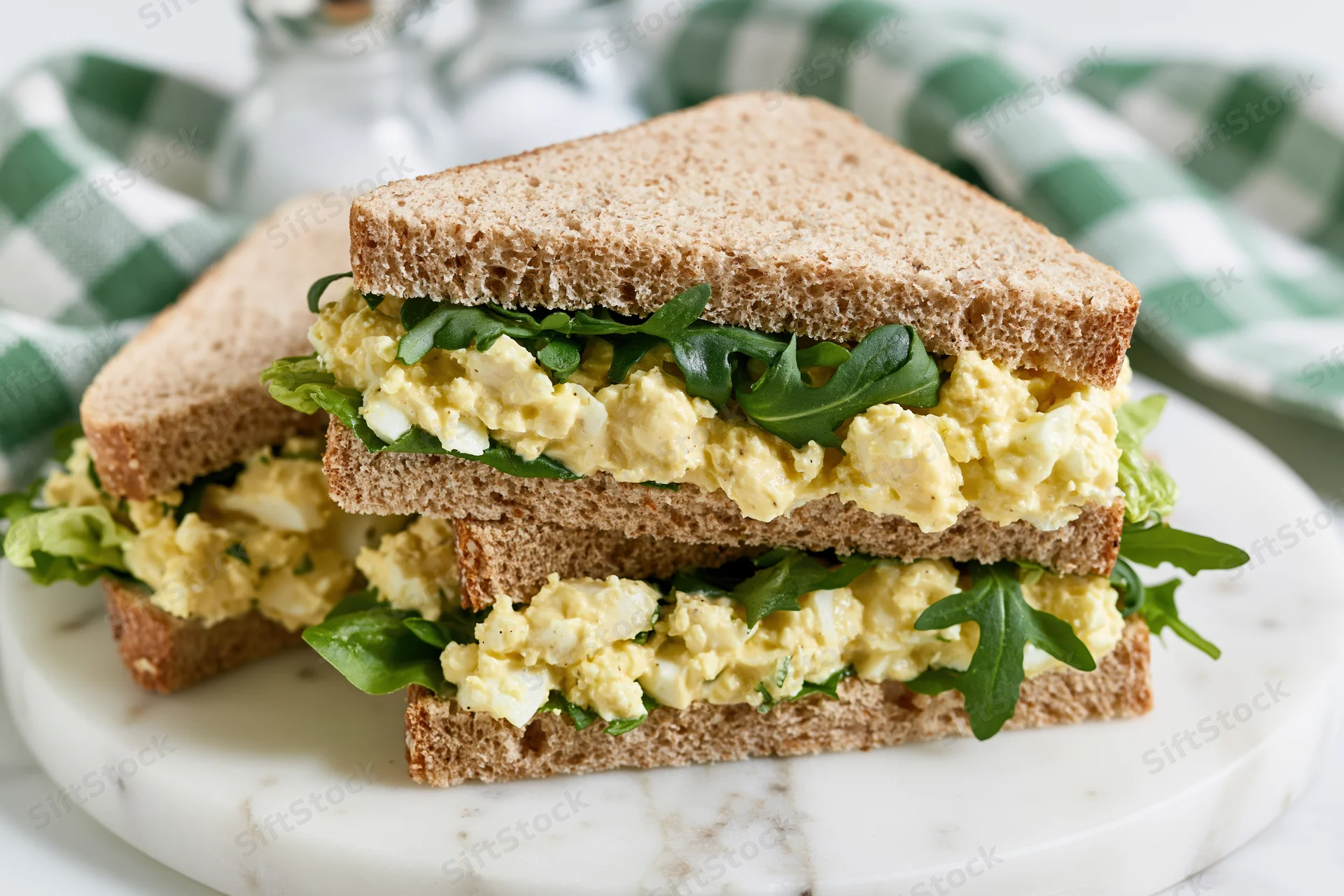 Egg salad sandwich cut in half and stacked on a white marble plate with arugula filling