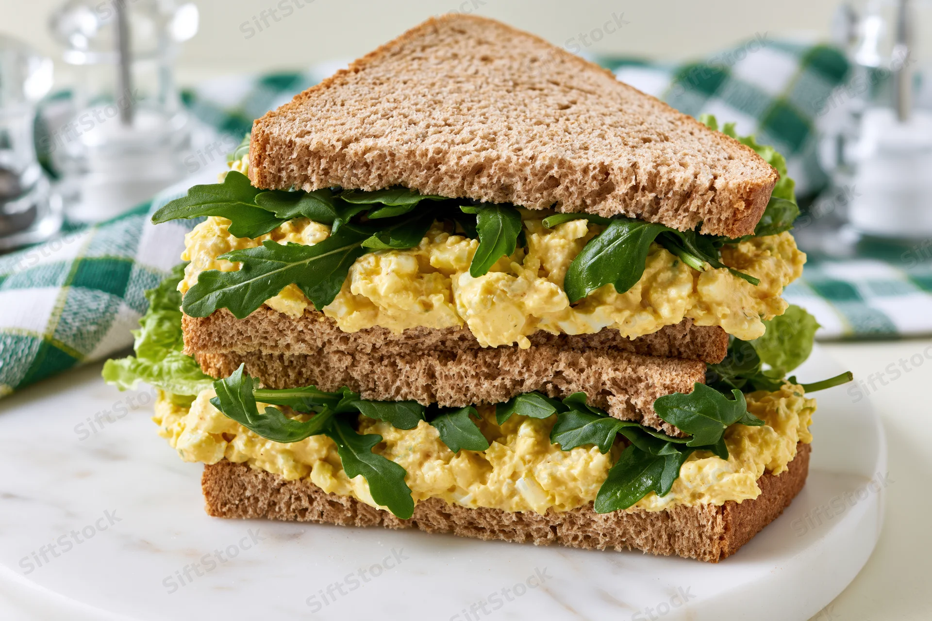 Egg salad sandwich cut in half and stacked on a white marble plate with arugula and lettuce layers