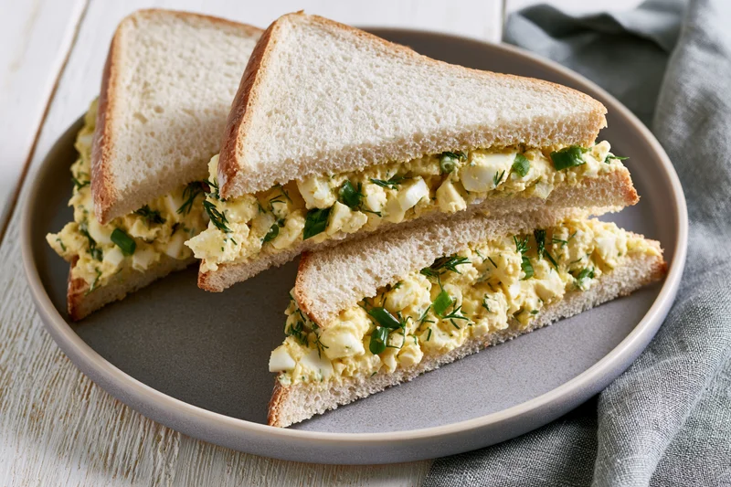 Egg salad sandwich cut into two triangles on a round grey plate with a cloth napkin beside it