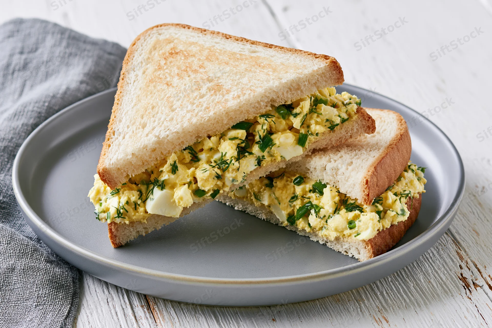 Egg salad sandwich cut into two triangles on a round grey plate with a wooden surface background