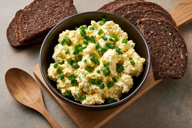 Egg salad with chopped green onions in a dark ceramic bowl served with rye bread slices on a wooden board