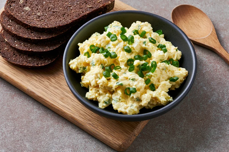 Egg salad with chopped green onions in a dark ceramic bowl on a wooden cutting board beside rye bread slices and a wooden spoon