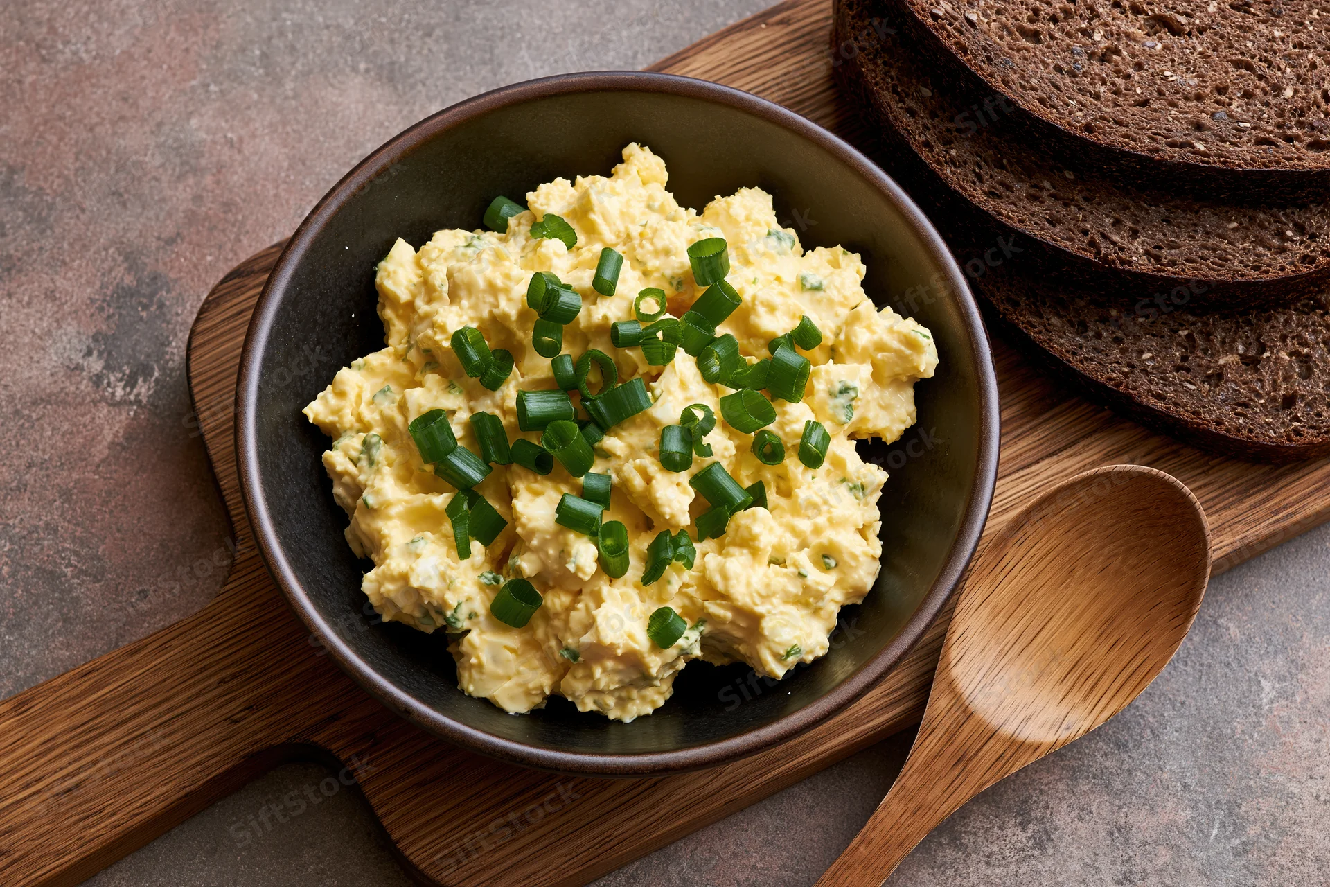 Egg salad with chopped green onions in a dark ceramic bowl on a wooden cutting board with rye bread slices and a wooden spoon beside it