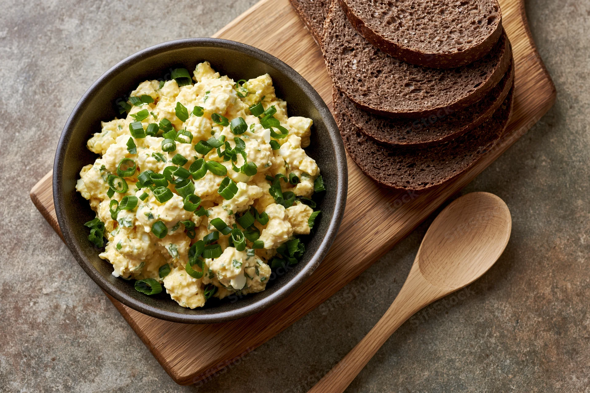 Egg salad with chopped green onions in a dark ceramic bowl on a wooden cutting board with rye bread slices and a wooden spoon beside it