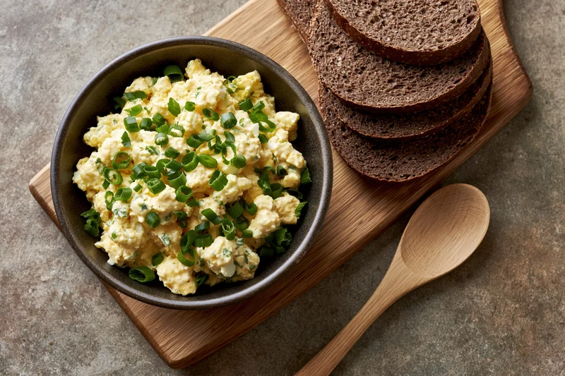 Egg salad with chopped green onions in a dark ceramic bowl on a wooden cutting board with rye bread slices and a wooden spoon beside it