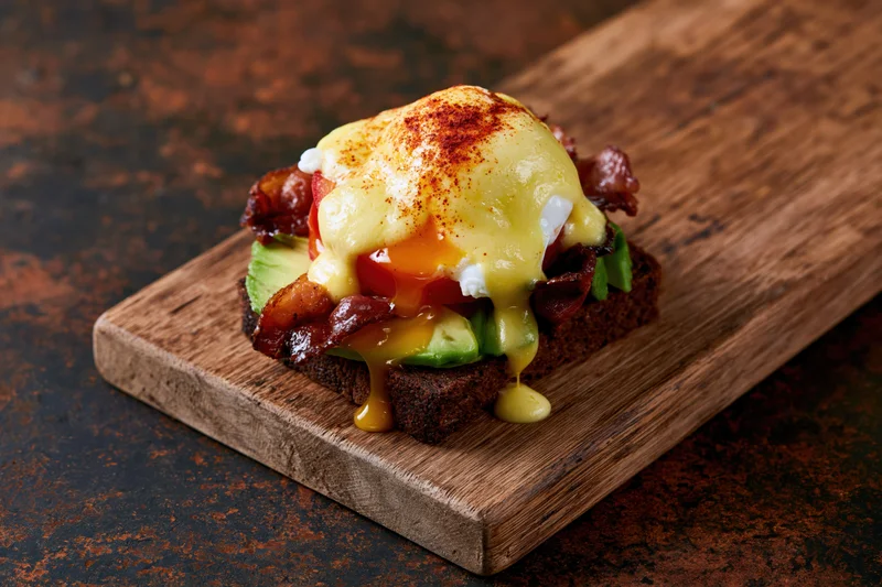 Open eggs benedict sandwich on dark rye toast with avocado and bacon, served on wooden board