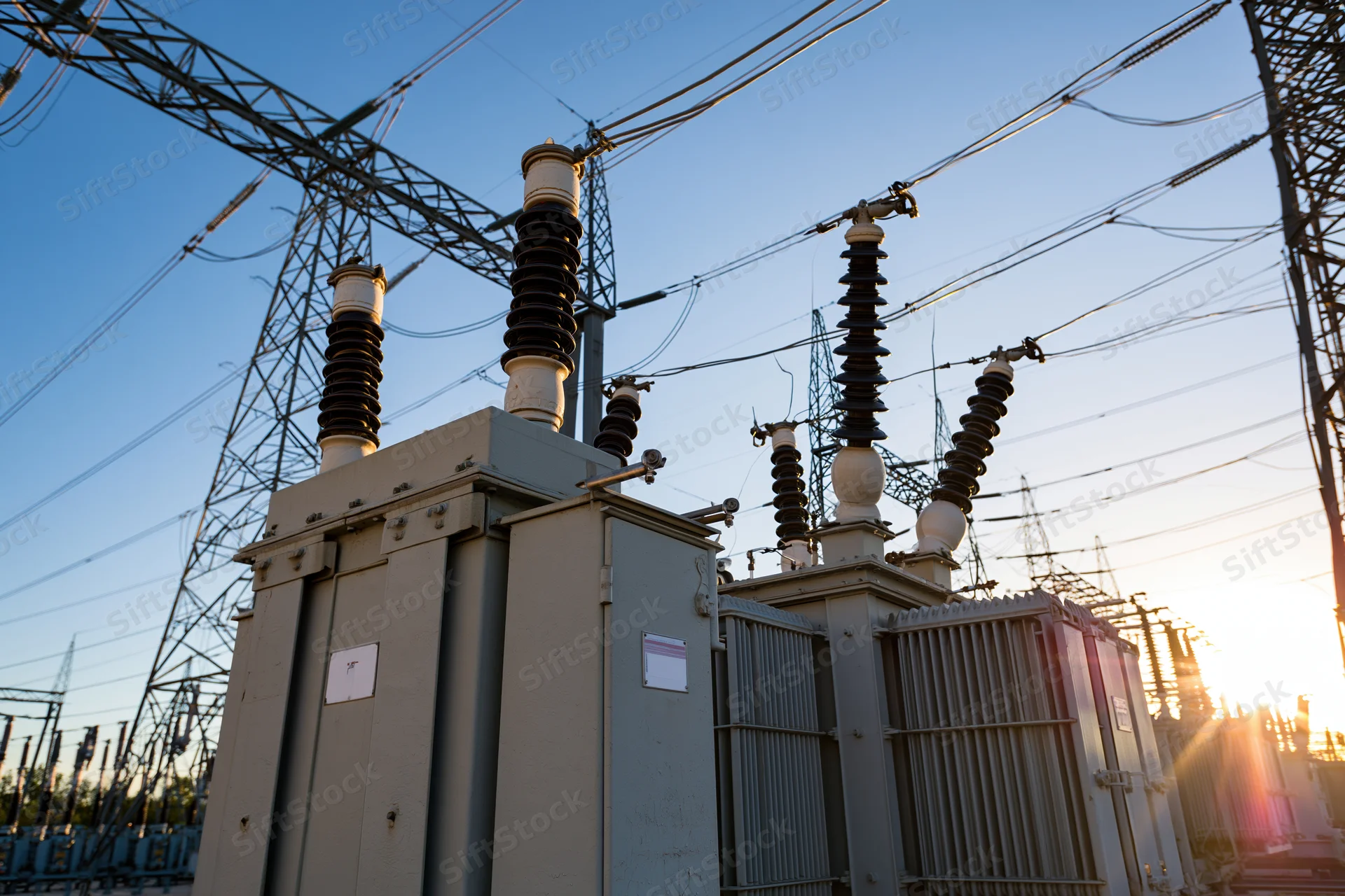 Electrical substation with transformers and transmission towers viewed from ground level at sunset