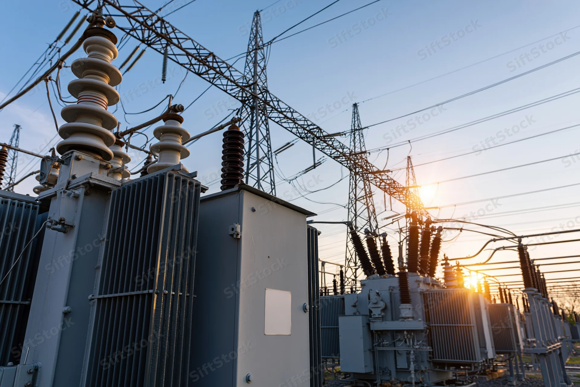 Electrical substation with transformers and transmission towers viewed from ground level at sunset
