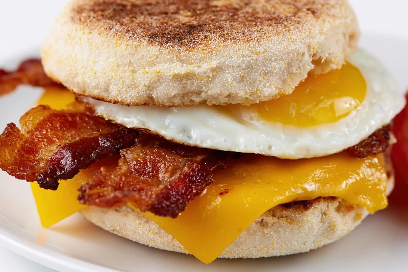 English muffin breakfast sandwich with fried egg, crispy bacon, and melted cheese on a white plate