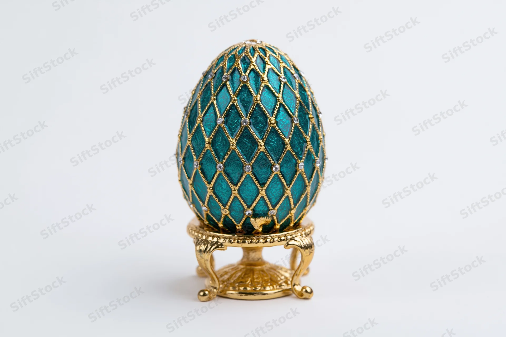 Teal enamel decorative egg with gold lattice and gemstones on ornate stand isolated on white