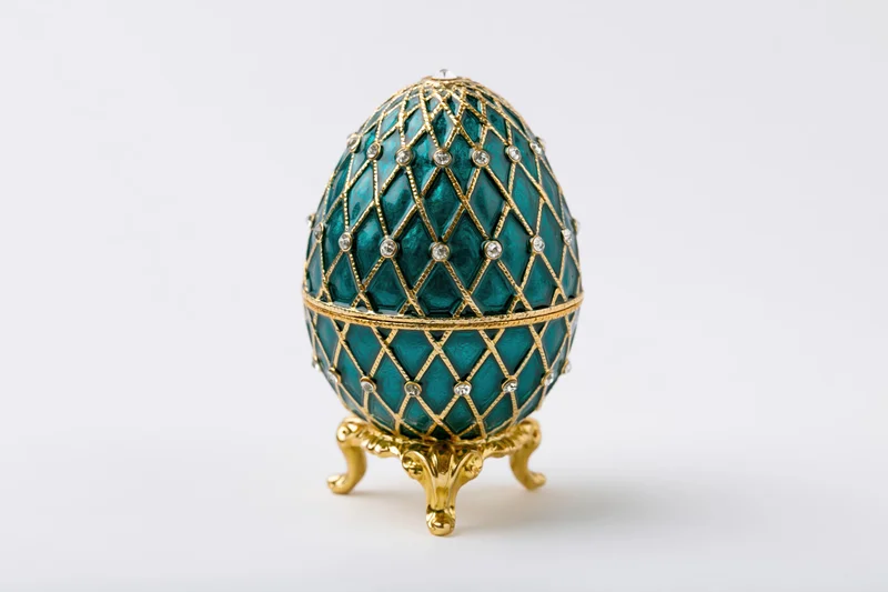 Teal enamel decorative egg with gold filigree and gemstones on a golden stand isolated on white