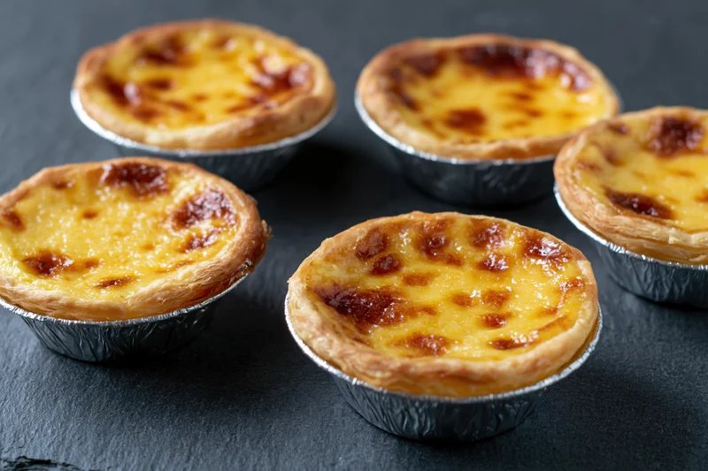 Five portuguese pastel de nata egg tarts in silver foil cups arranged on dark surface