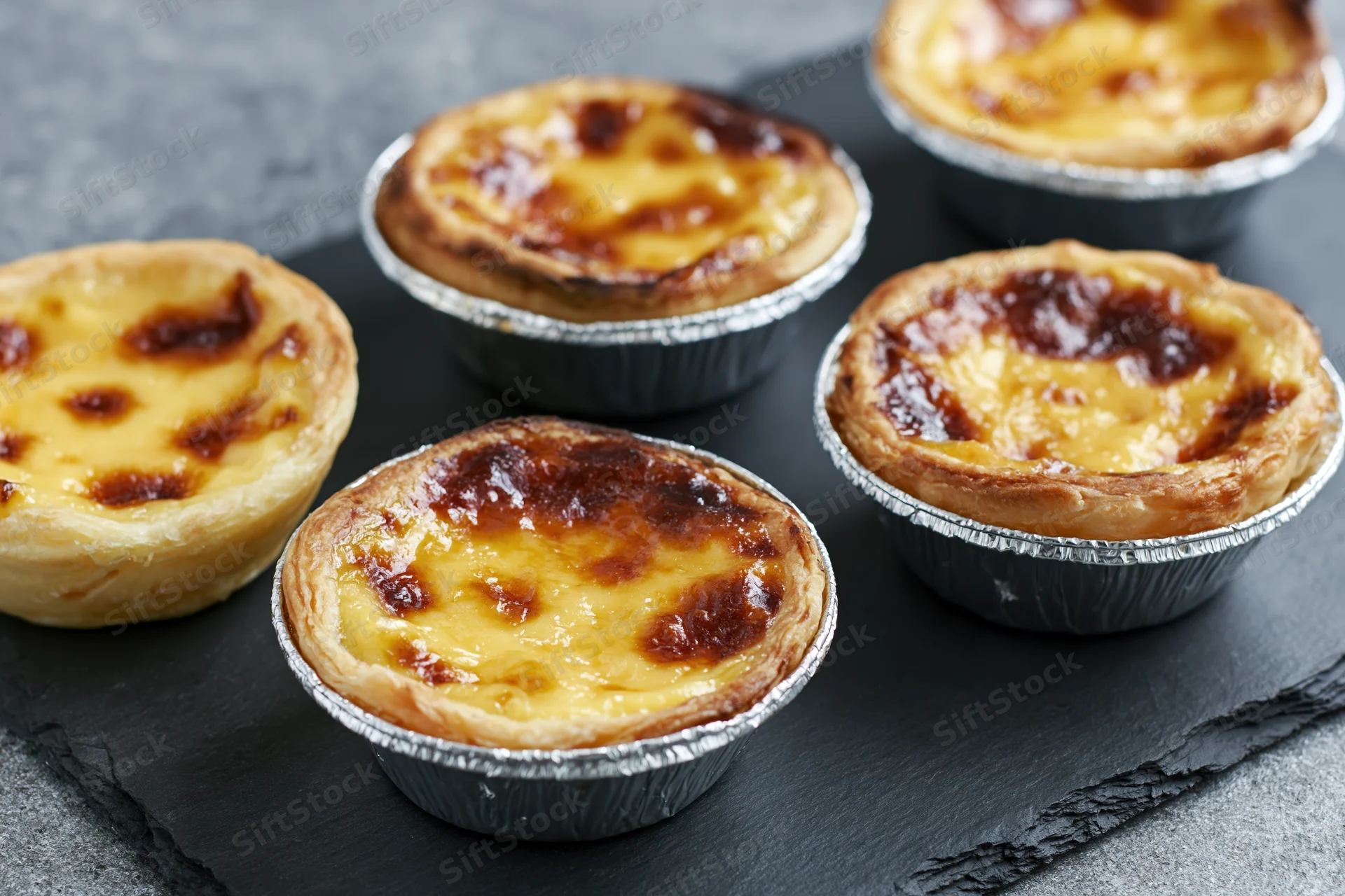 Five portuguese pastel de nata egg tarts in silver foil cups arranged on a black slate tray