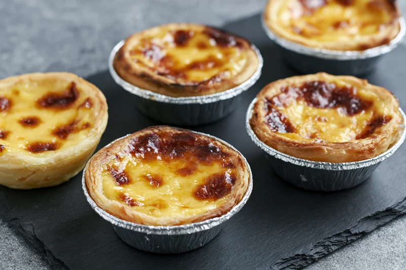 Five portuguese pastel de nata egg tarts in silver foil cups arranged on a black slate tray
