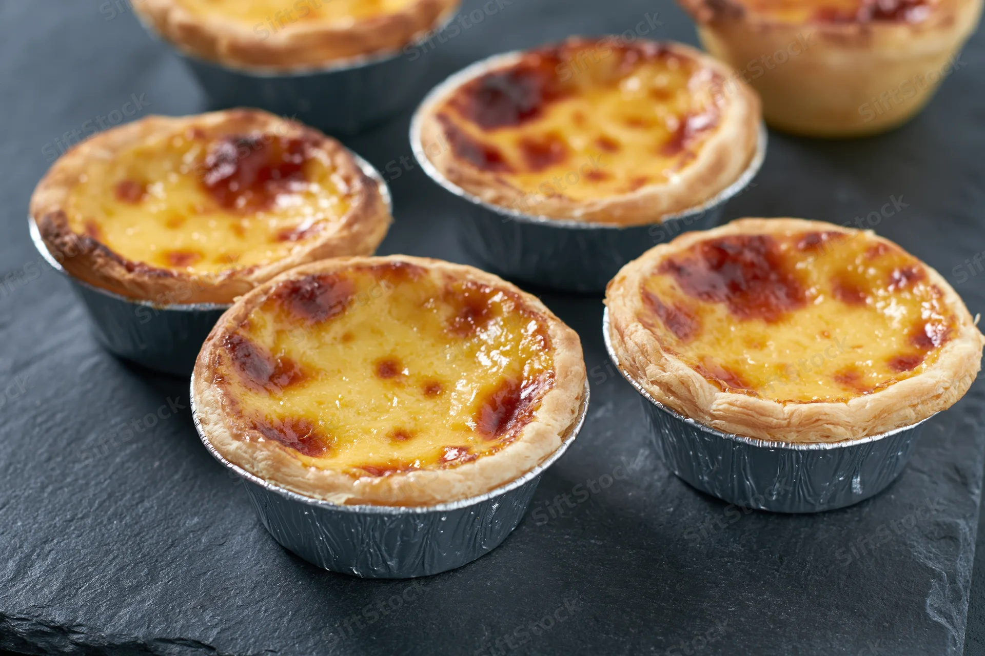 Five portuguese pastel de nata egg tarts in silver foil cups arranged on a dark slate surface