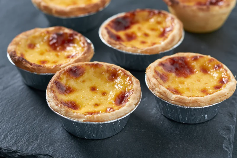 Five portuguese pastel de nata egg tarts in silver foil cups arranged on a dark slate surface