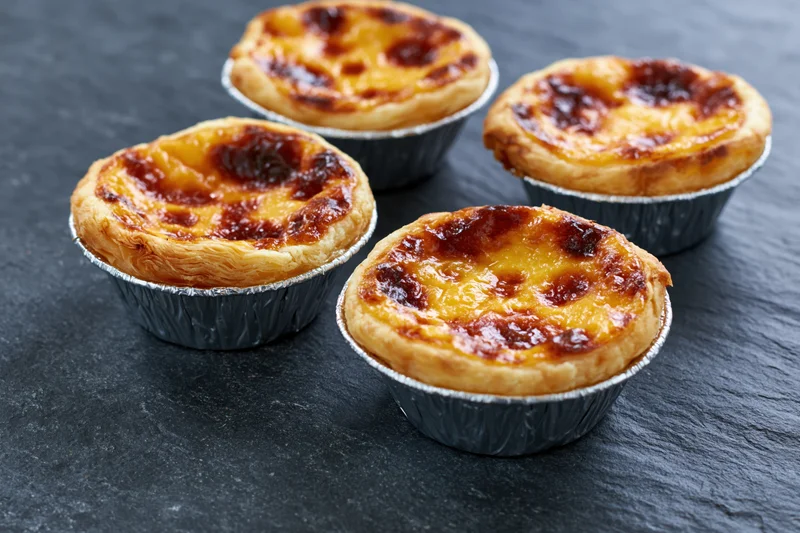 Four pastel de nata egg tarts in silver foil cups arranged on a dark slate surface