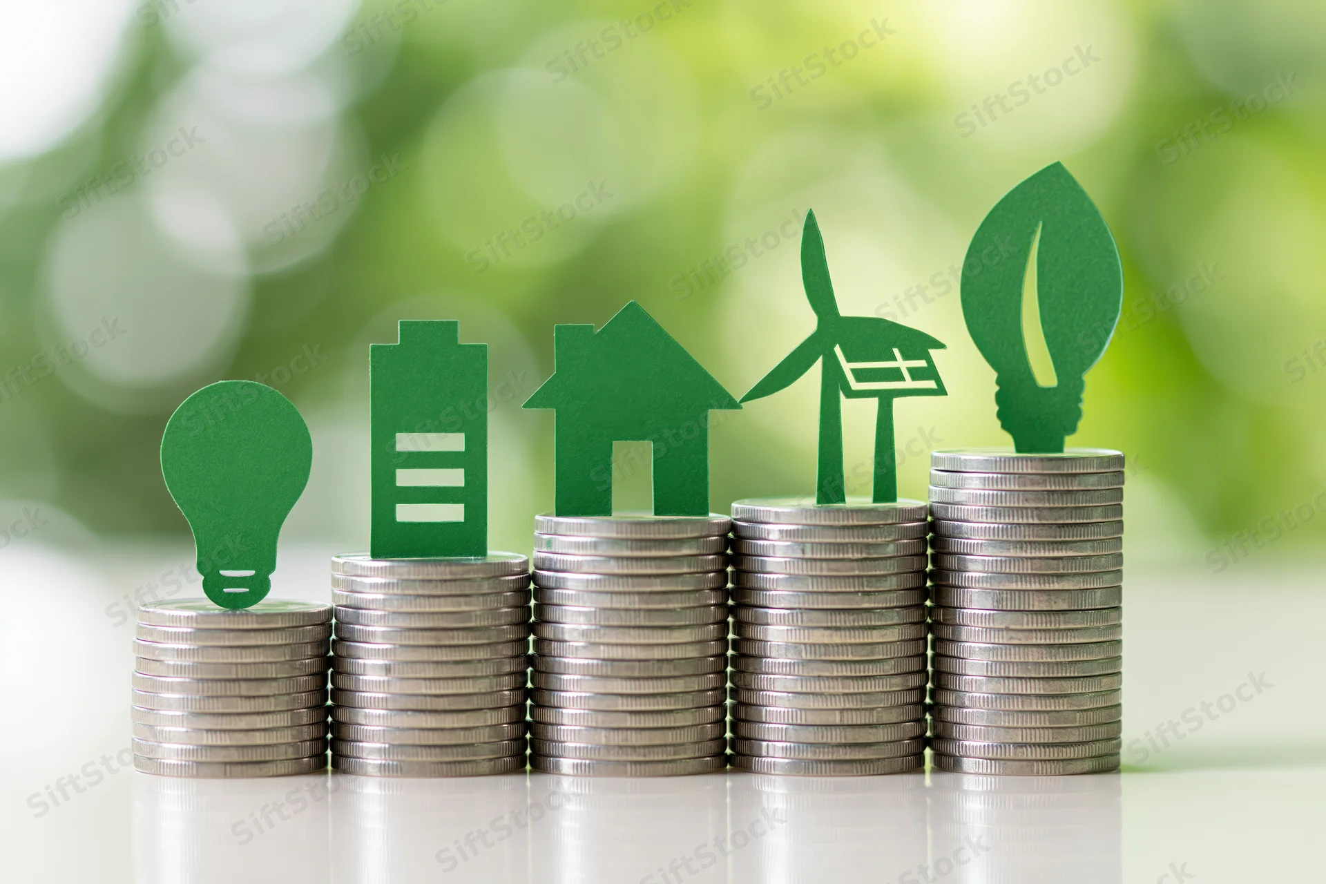 Five ascending stacks of silver coins each topped with green paper cutouts of lightbulb, building, house, wind turbine, and leaf on a reflective surface with blurred green background