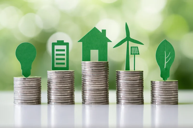 Five stacks of silver coins with green paper cutouts of energy symbols arranged in ascending height on a reflective surface with blurred green background