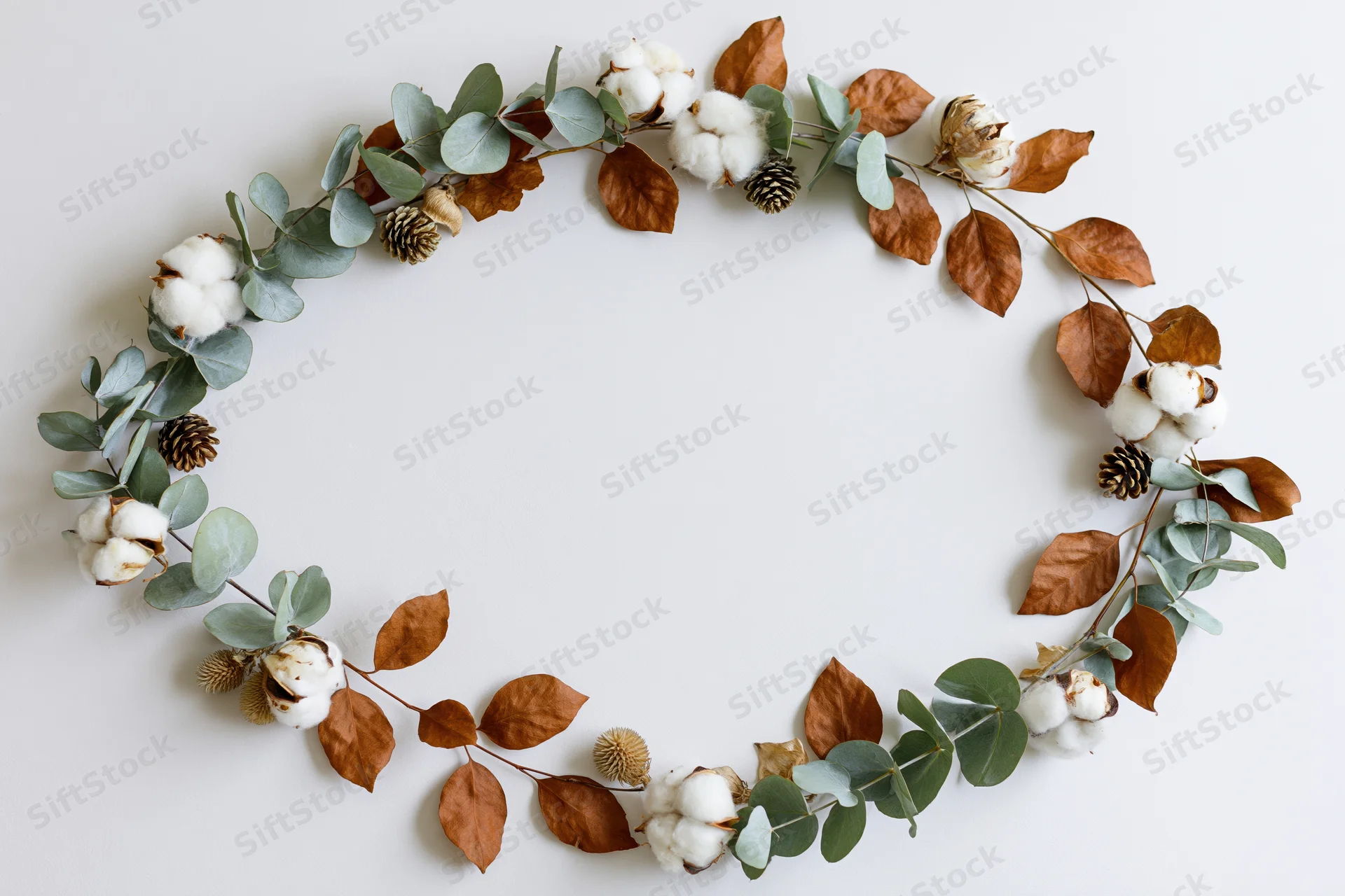 Circular wreath made of eucalyptus branches, dried cotton bolls, and brown leaves arranged flat on white surface
