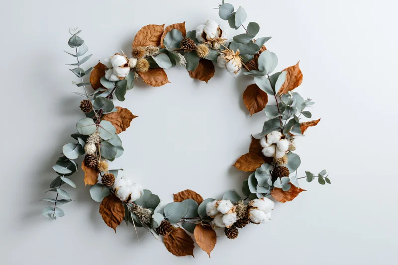 Circular wreath made of eucalyptus, dried cotton, pinecones and brown leaves arranged flat on white surface