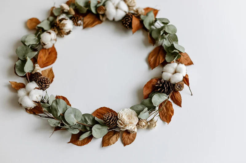 Circular wreath made of eucalyptus, dried cotton bolls, pinecones and autumn leaves arranged flat on white surface