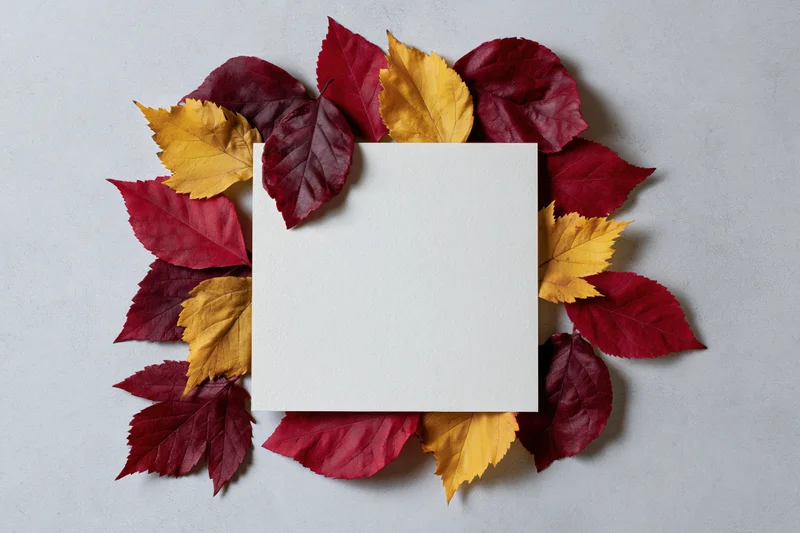 Blank white square card centered on a flat lay with autumn leaves arranged around it on a light surface
