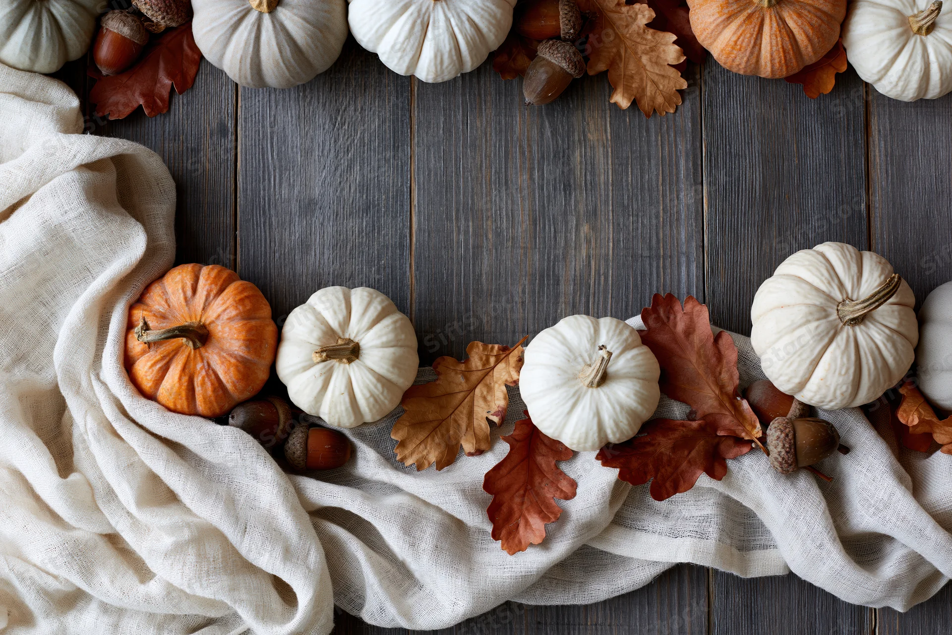 Small decorative pumpkins and dried oak leaves arranged in a flat lay on a wooden table with linen fabric and acorns forming a border with copyspace in the center