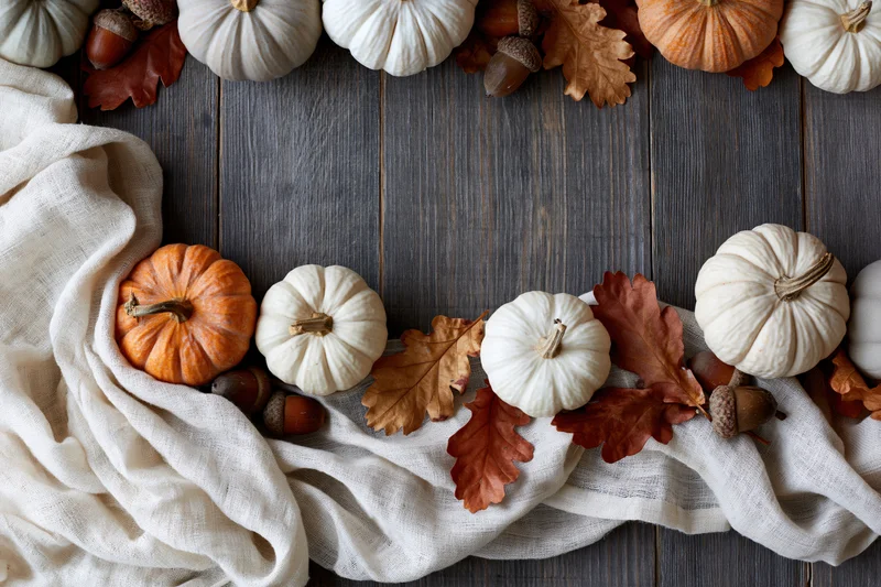 Small decorative pumpkins and dried oak leaves arranged in a flat lay on a wooden table with linen fabric and acorns forming a border with copyspace in the center