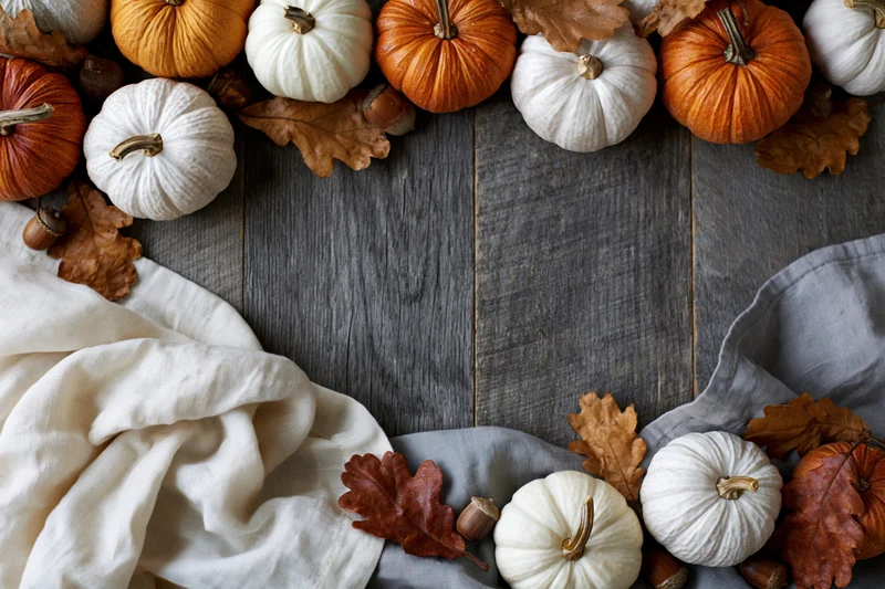 Small decorative pumpkins and dried oak leaves arranged in a border on a wooden surface with fabric and acorns