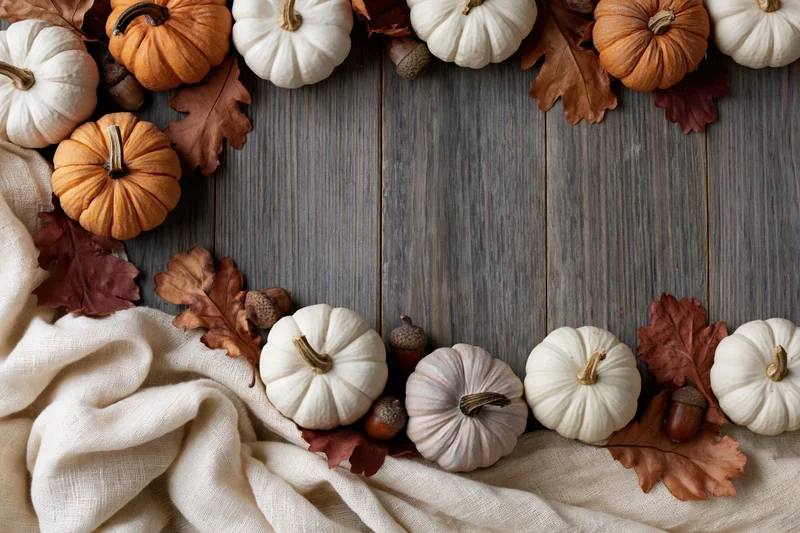 Small decorative pumpkins and dried oak leaves arranged in a border on a wooden table with fabric and acorns