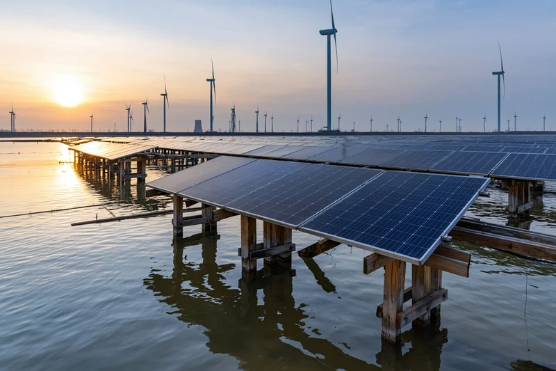 Floating solar panels mounted on wooden stilts over calm water with wind turbines in the distance at sunset